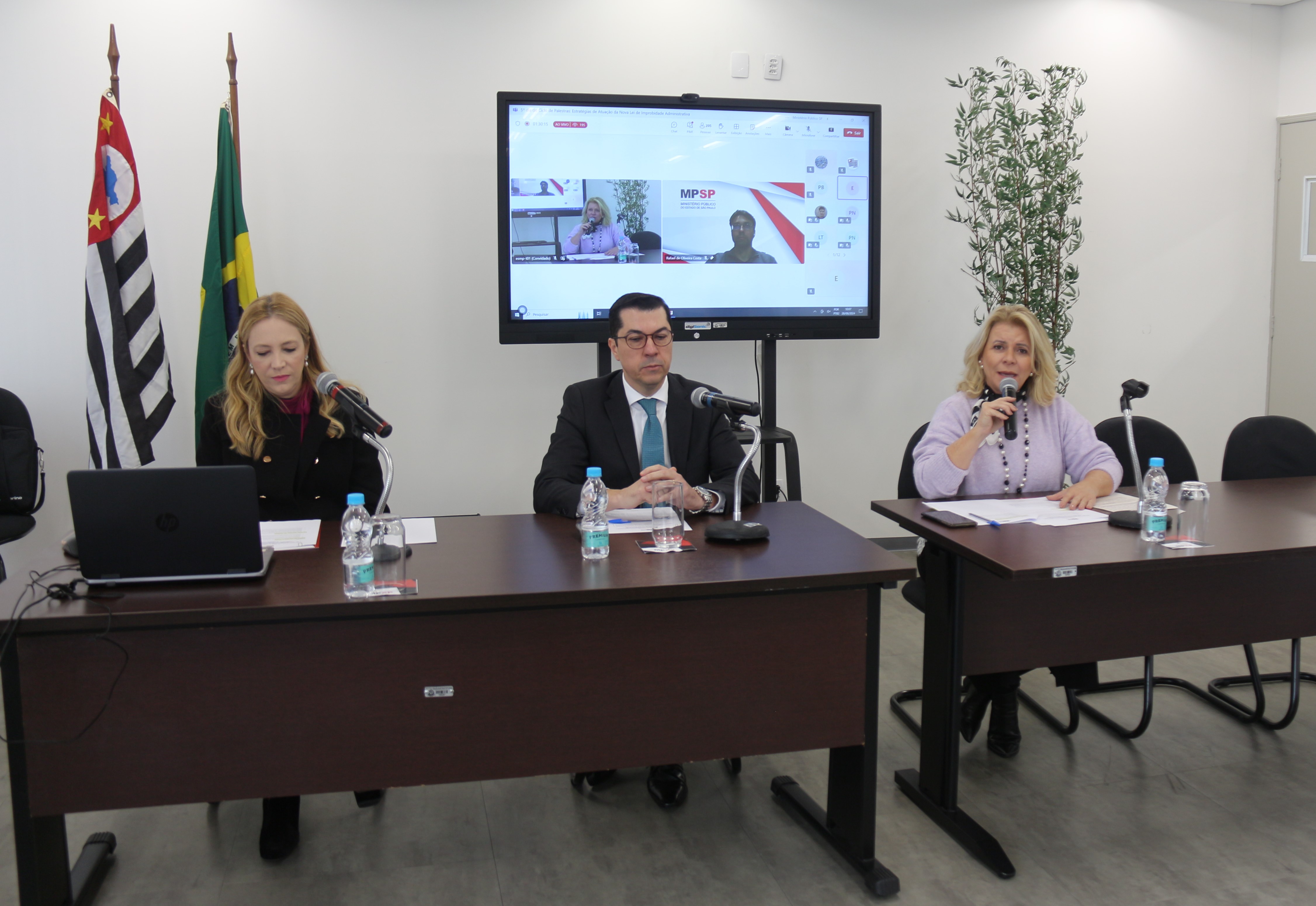 Foto mostra a promotora de Justiça e mediadora, o procurador da República e palestrante e a procuradora de Justiça e assessora da ESMPSP. A assessora fala ao microfone. Todos estão sentados à mesa de debates. Há copos e garrafinhas de água sobre as mesas de cor marrom. Há um telão ao fundo, sobre uma parede branca, em que aparecem a assessora e o segundo palestrante, que participou remotamente. Ao lado de uma das mesas, estão as bandeiras do Brasil e de São Paulo. Fim da descrição.  