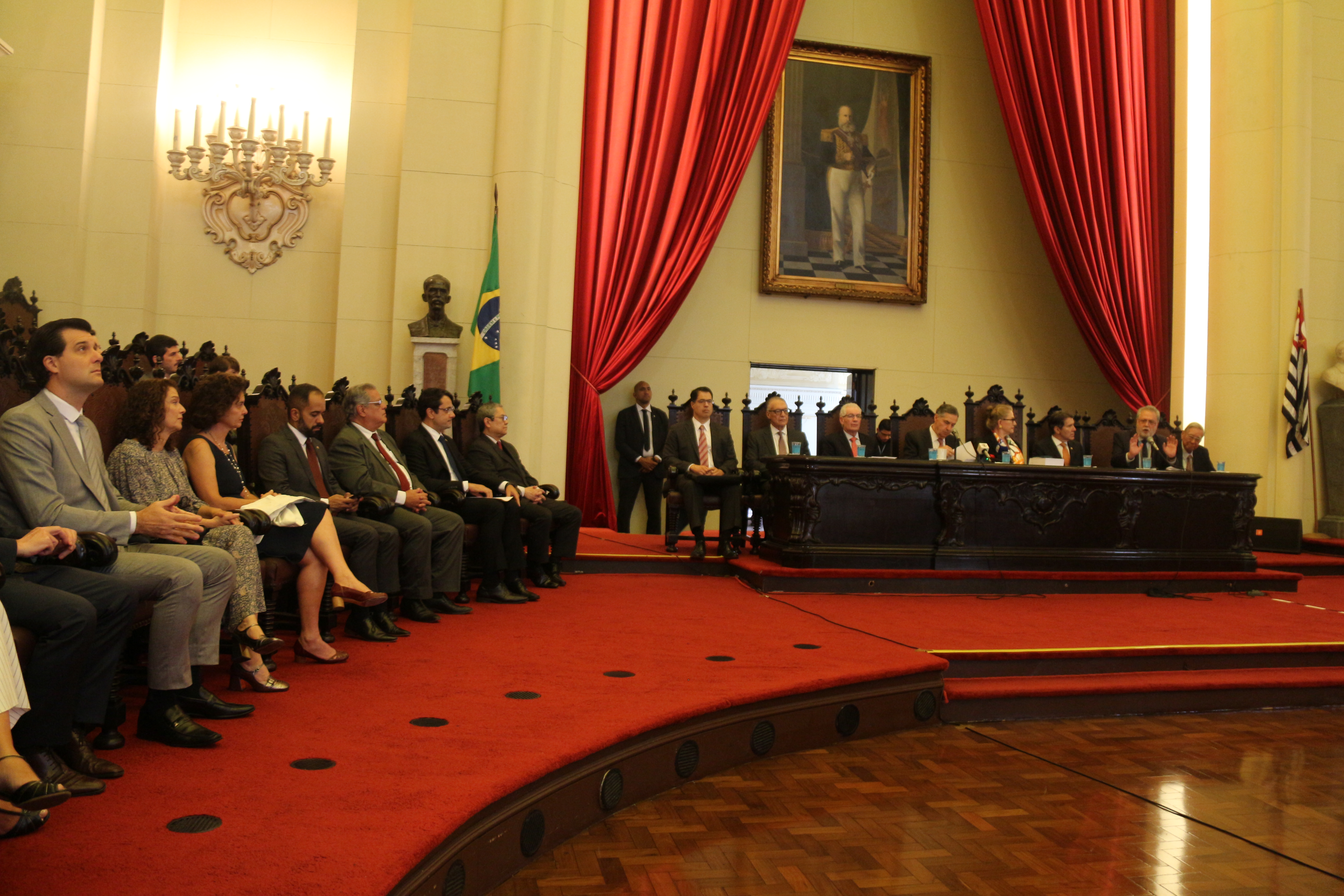 Foto mostra o salão de honra, com a mesa principal entre cortinas vermelhas e um quadro grande na parede. O carpete também é vermelho.  Entre os participantes, está o PGJ e outras autoridades. Na mesa lateral, estão a diretora da ESMPSP e demais convidados de honra. Na parede ao fundo, há um lustre grande, um busto e a bandeira do Brasil. Do outro lado da mesa, está a bandeira de São Paulo. No rodapé, à direita, está o logo do MPSP em preto e vermelho. Fim da descrição. 