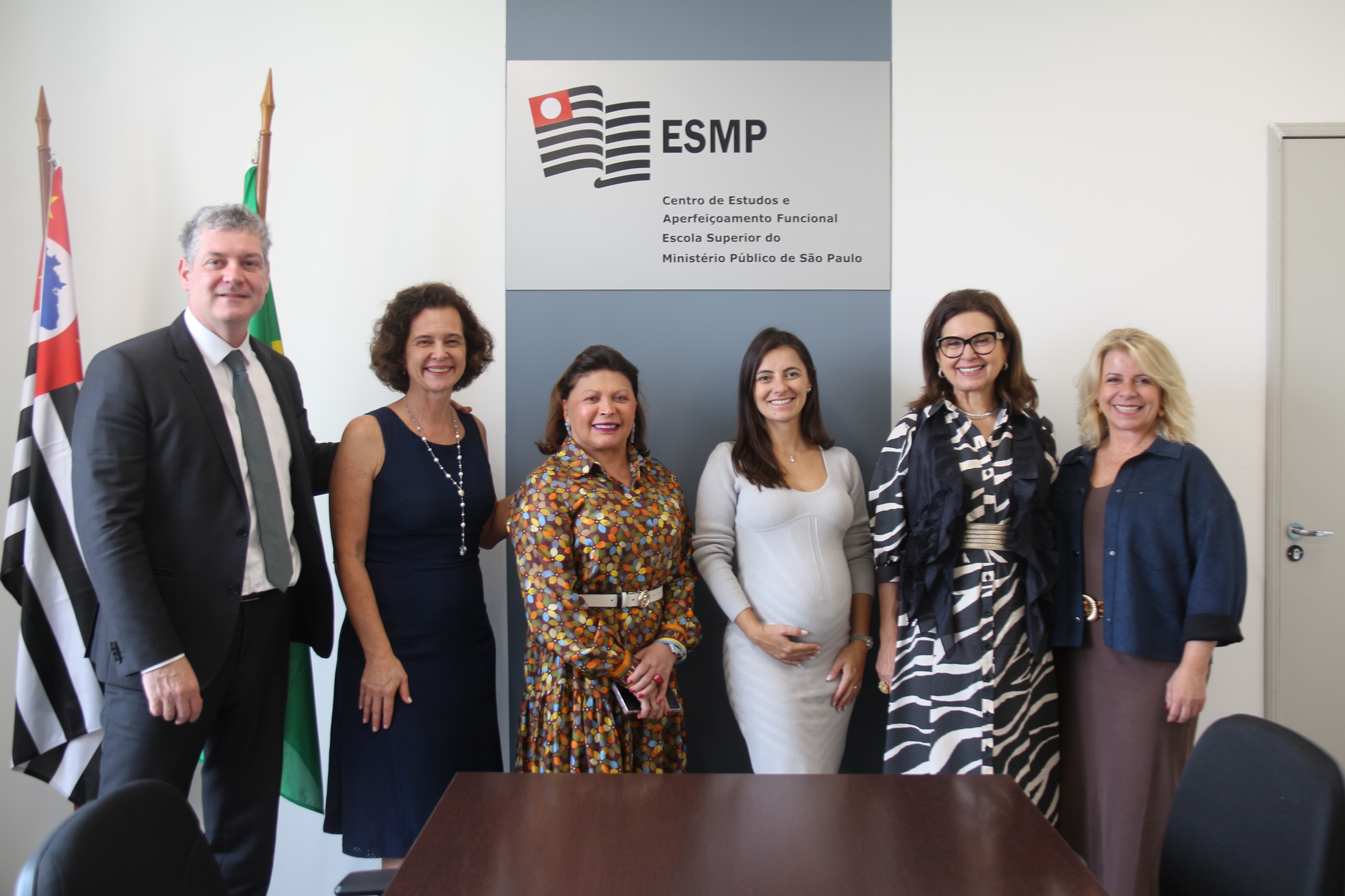 Foto mostra a diretora da ESMPSP e dois de seus assessores ao lado da vice-diretora e da diretora financeira do CDEMP e da promotora de Justiça do MPPR. Eles estão de pé e sorrindo em frente à placa do CEAF/ESMP, com o logo em preto e vermelho sobre uma parede branca. Atrás do assessor, estão as bandeiras do Brasil e de São Paulo. Fim da descrição.