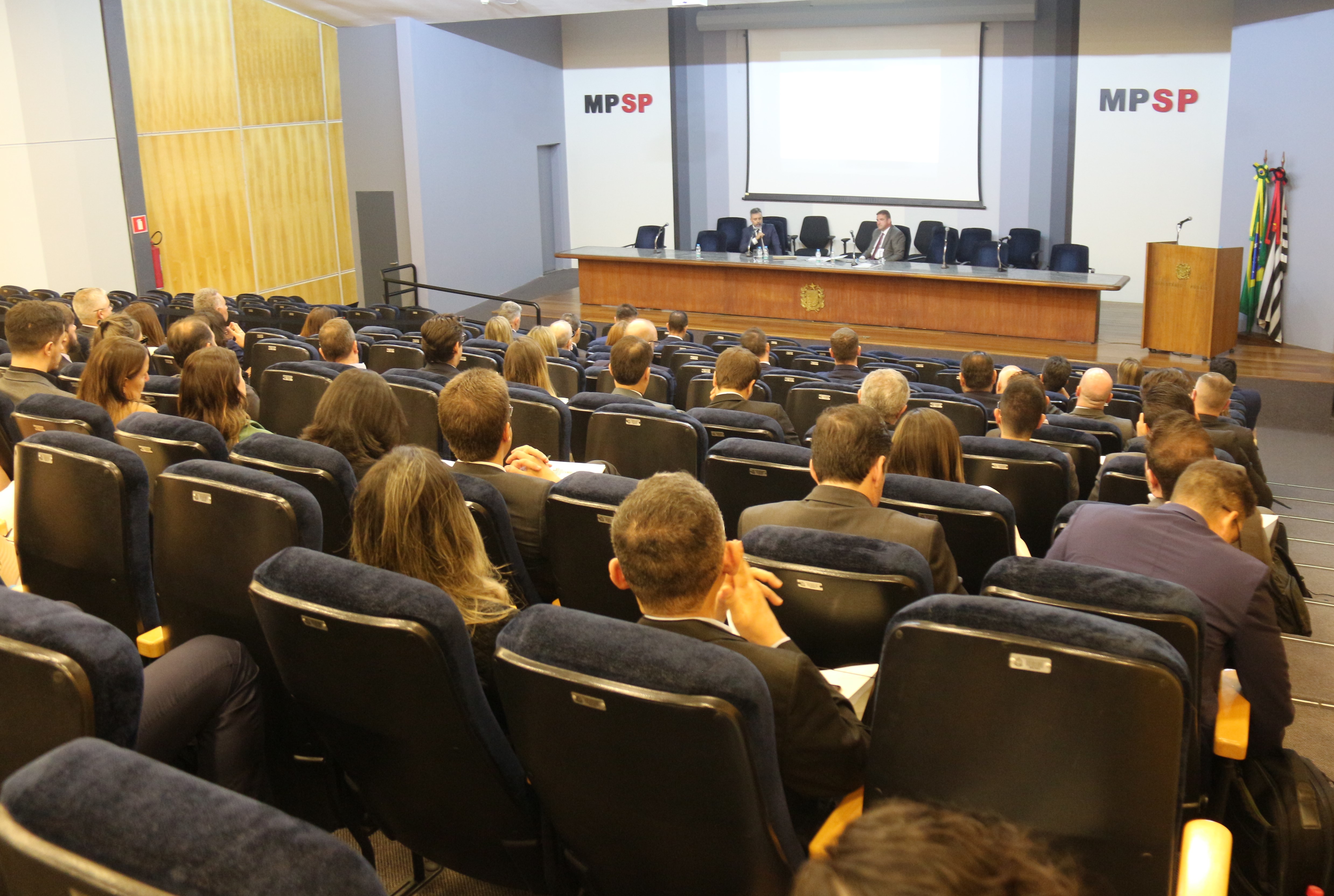 Foto mostra mesa de debates (com dois expositores) e plateia vistas do alto. As poltronas são pretas, a mesa e o púlpito são marrons. Ao fundo, há um telão de projeção e a sigla do MPSP em preto e vermelho. Ao lado esquerdo da mesa, estão as bandeiras do Brasil e de São Paulo. Na parede à esquerda da plateia, há um extintor de incêndio.  Fim da descrição.