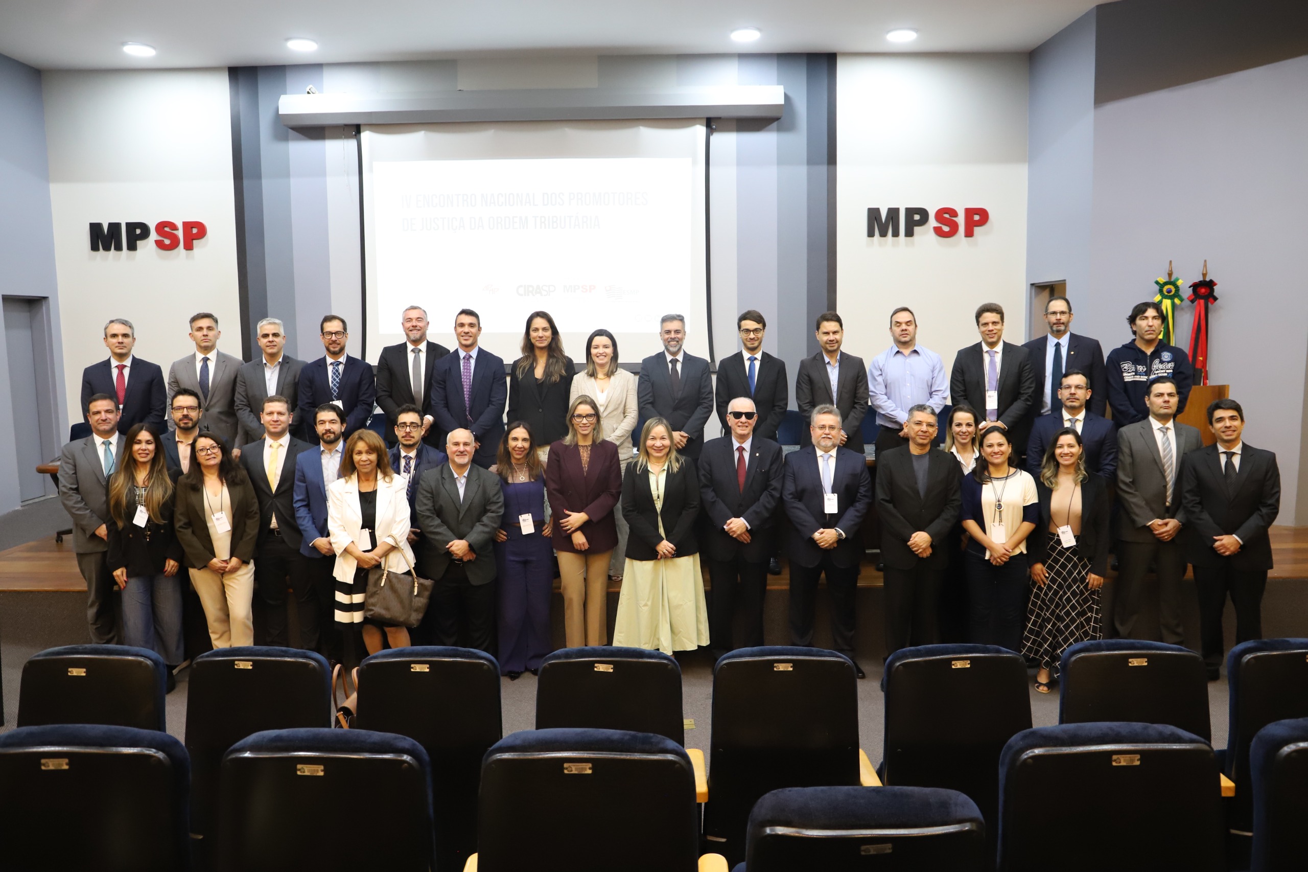 foto mostra os(as) participantes do evento em frente à mesa de debates do auditório Queiroz Filho. Todos estão de pé. Na parede branca, ao fundo, está o logo do MPSP em preto e vermelho dos lados direito e esquerdo do telão de projeção. Fim da descrição.