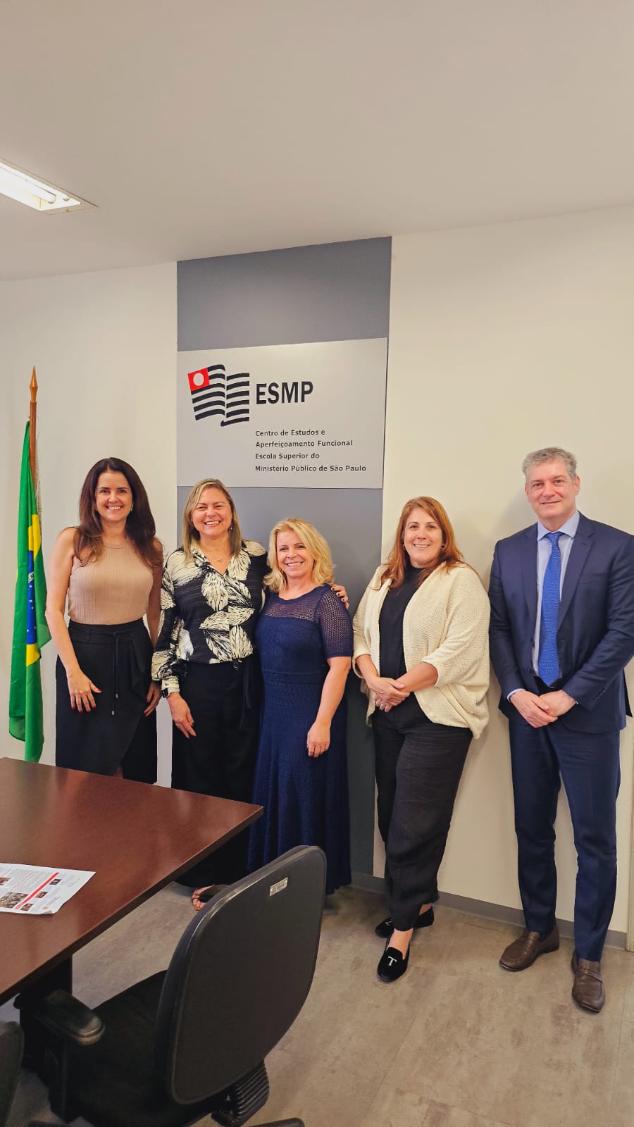 foto mostra a promotora de Justiça e chefe de gabinete do MPSP e os promotores de Justiça e assessores da ESMPSP ao lado da assessora técnico-pedagógica do CEAF/RN.  Eles estão de pé e sorrindo em frente à placa da ESMP. Ao lado de uma das assessoras, está a bandeira do Brasil. Fim da descrição. 