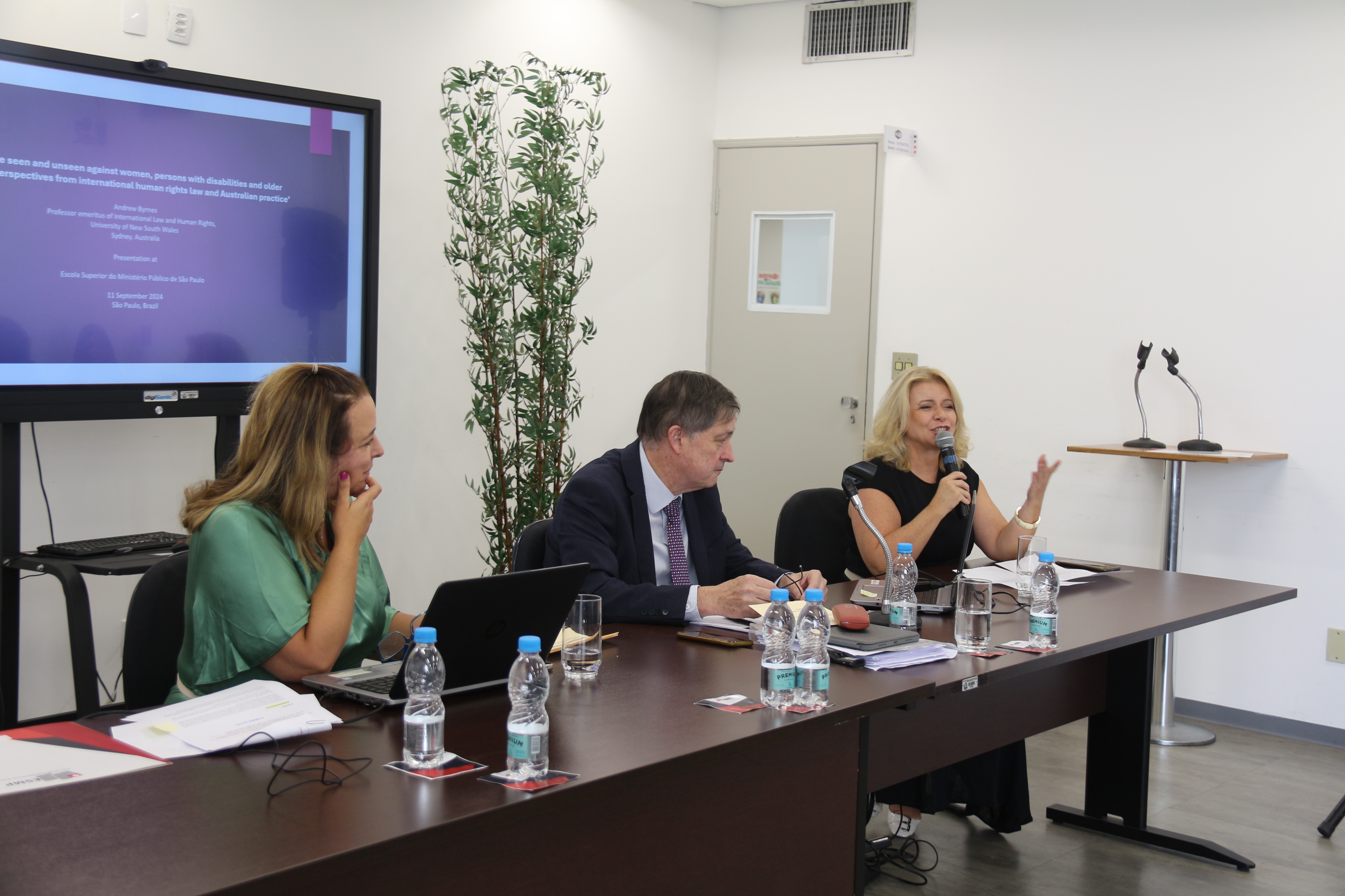 Foto mostra a assessora da ESMPSP falando ao microfone. Ela está ao lado do palestrante e da tradutora na mesa de debates. As duas promotoras de Justiça são mulheres brancas e têm cabelos castanhos claros. O professor australiano é homem branco e tem cabelos castanhos escuros. Há garrafas e copos de água sobre a mesa marrom. É possível ver a porta lateral e o ar-condicionado um pouco acima. Fim da descrição. 