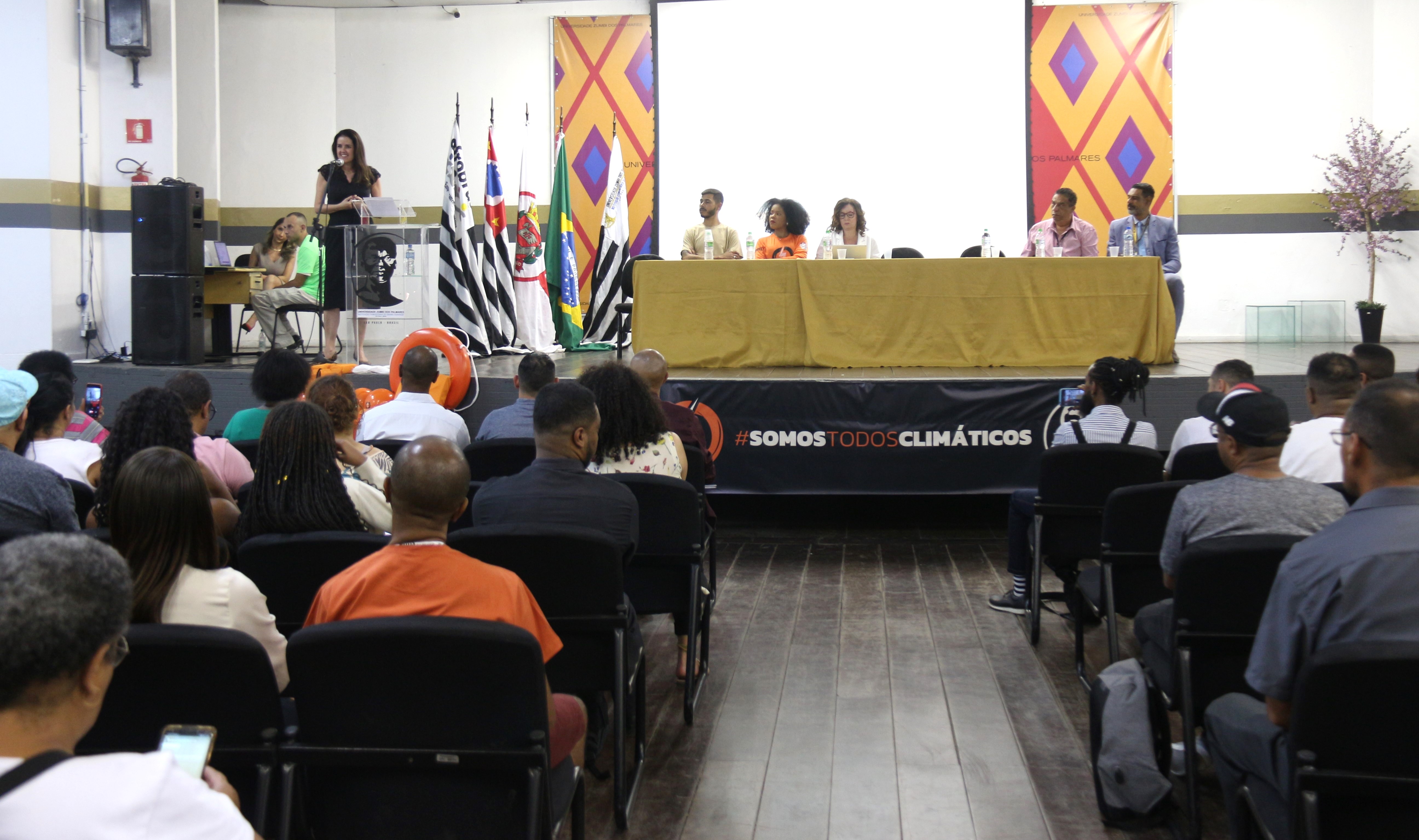 A promotora Florenci Milani discursa à frente da plateia que está no auditório. Ela está de pé e fala ao microfone em frente ao púlpito. Ao lado, na mesa de debates, estão sentados cinco participantes, três homens e duas mulheres. À frente da mesa, está escrito Somos todos climáticos. Fim da descrição. 