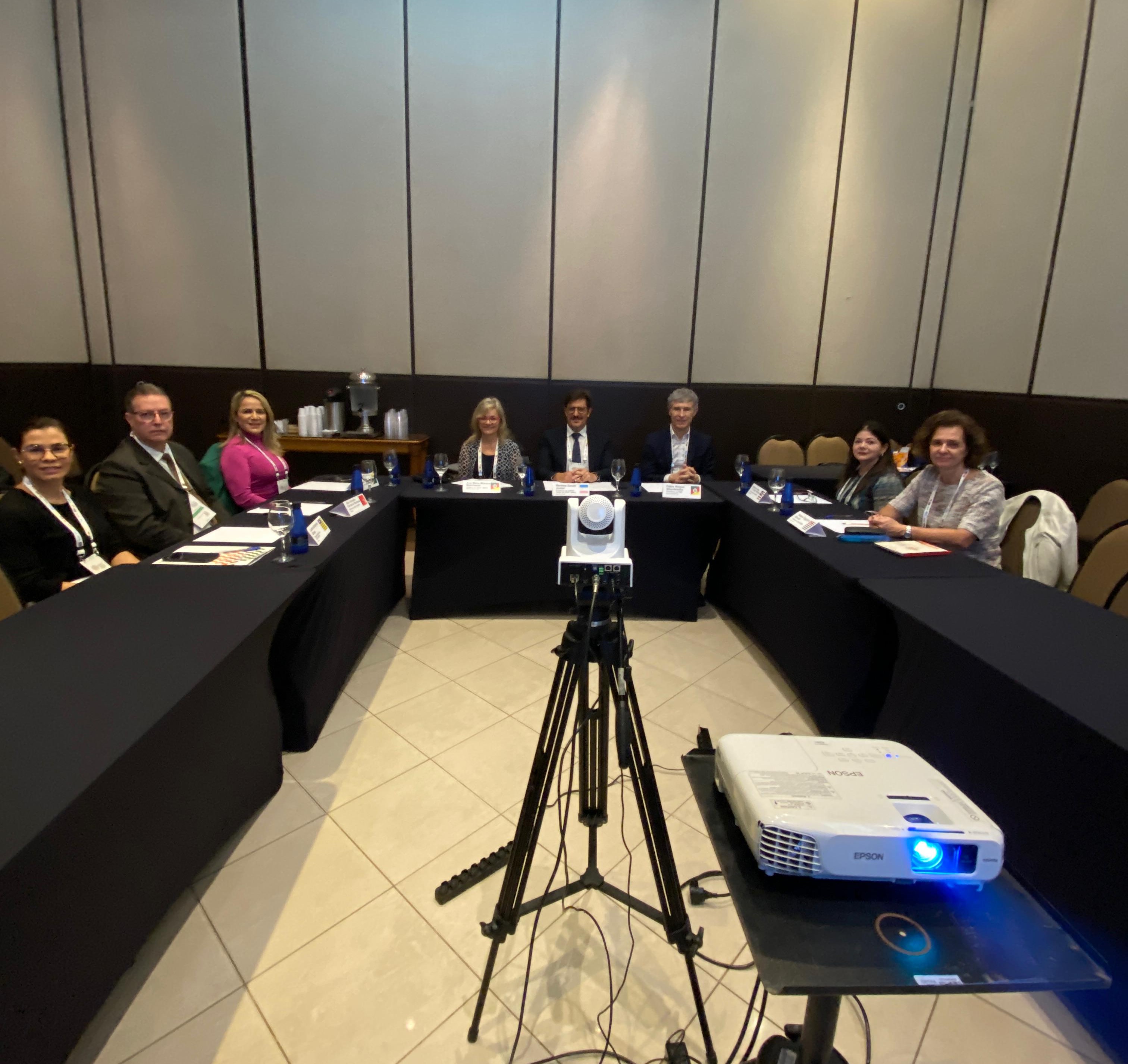 foto mostra a diretora da ESMPSP e alguns participantes às mesas de debates. À frente da sala, há uma câmera com tripé e um projetor de slides sobre um aparador. Fim da descrição.