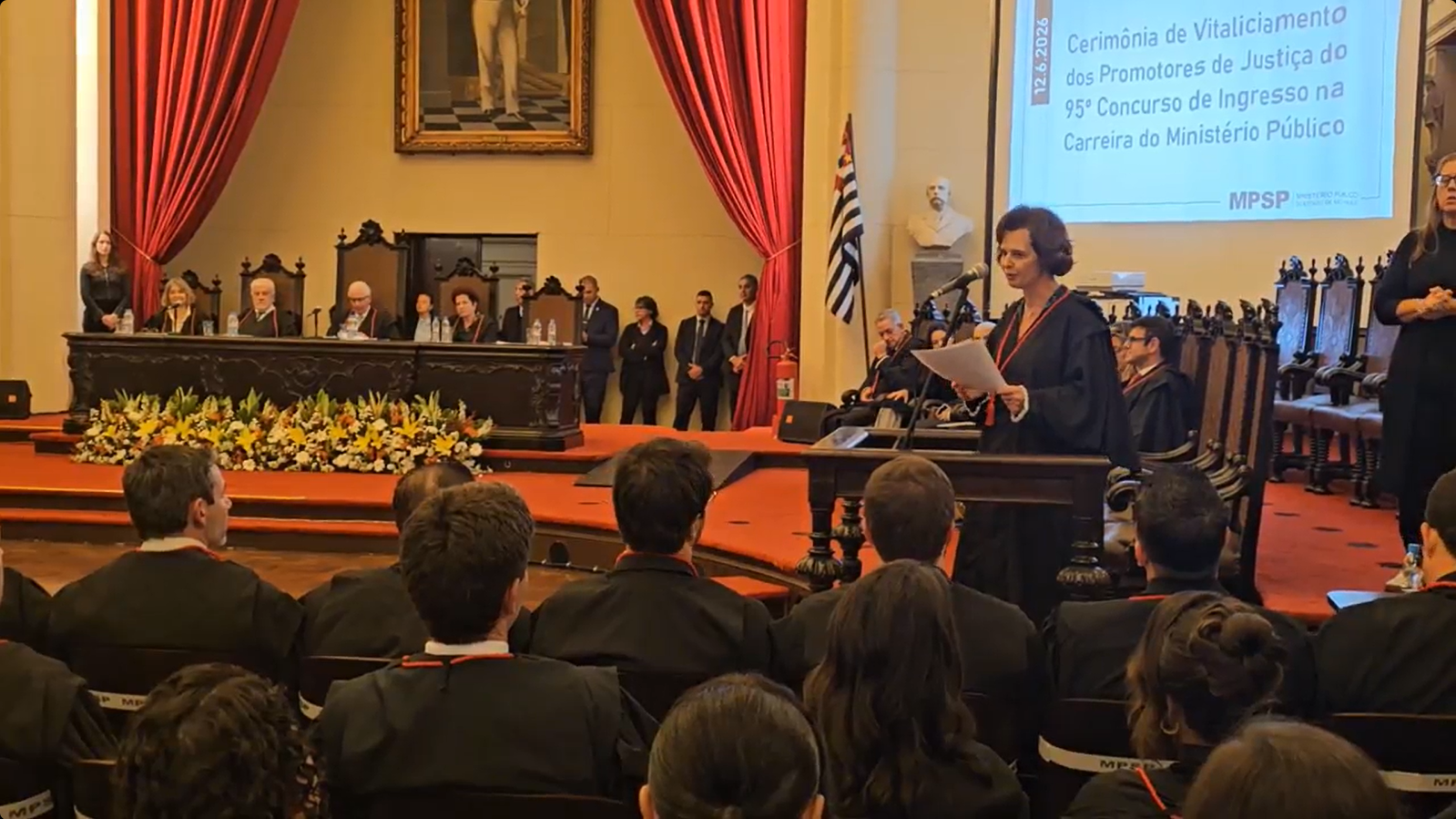 Foto mostra a diretora da ESMPSP discursando, a mesa de honra e parte da plateia. Fim da descrição.